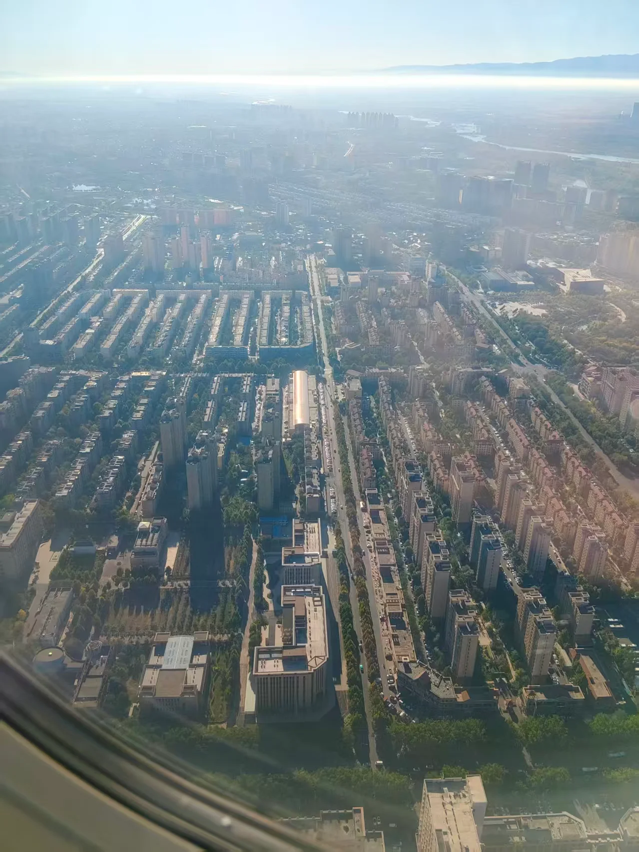 从飞机上俯拍城市，密集楼群、道路与大片绿地沿画面纵向延伸，远处天空和薄雾映出柔和光线。